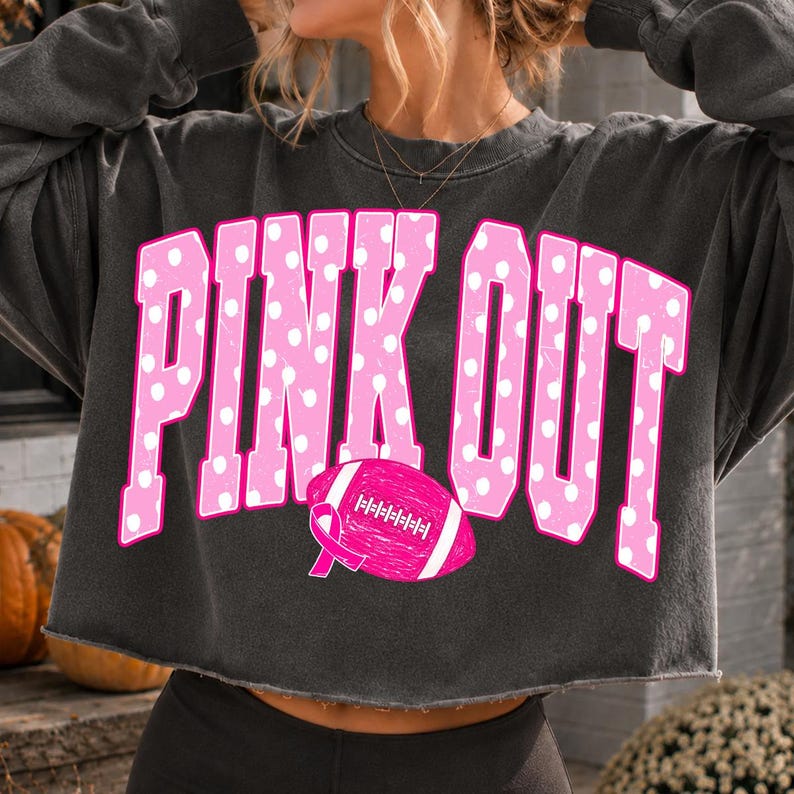 May include: A charcoal gray cropped sweatshirt featuring the text PINK OUT in large, distressed pink letters with white polka dots. A pink football graphic with a breast cancer awareness ribbon is positioned below the text on the front.