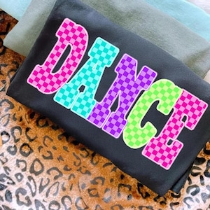 Puede incluir: Sudadera negra con la palabra "DANCE" en letras grandes y coloridas a cuadros. Las letras están delineadas en blanco y presentan cuadrados rosas, turquesas, morados y verdes. La sudadera está doblada sobre otra tela.