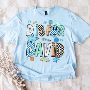 May include: A light blue t-shirt featuring a graphic print with the text DIS FOR DAVID. The design includes school-themed icons like a pencil, globe, apple, and school bus, alongside checkered, striped, and star patterns in blue, orange, and teal.