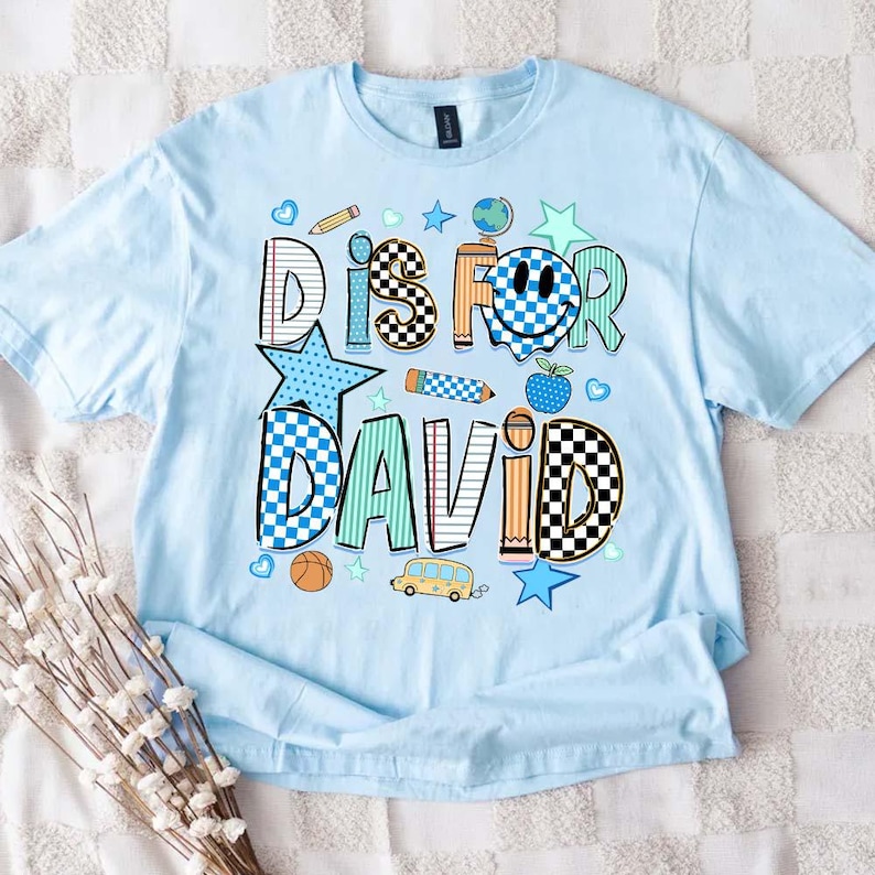 May include: A light blue t-shirt featuring a graphic print with the text DIS FOR DAVID. The design includes school-themed icons like a pencil, globe, apple, and school bus, alongside checkered, striped, and star patterns in blue, orange, and teal.