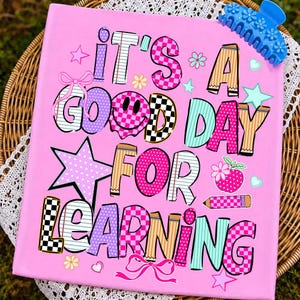 May include: A rectangular pink sign featuring the text IT'S A GOOD DAY FOR LEARNING in multi-coloured, patterned letters. The design includes a smiley face, stars, bows, a pencil, and an apple icon. A blue floral hair claw clip rests on the top right.