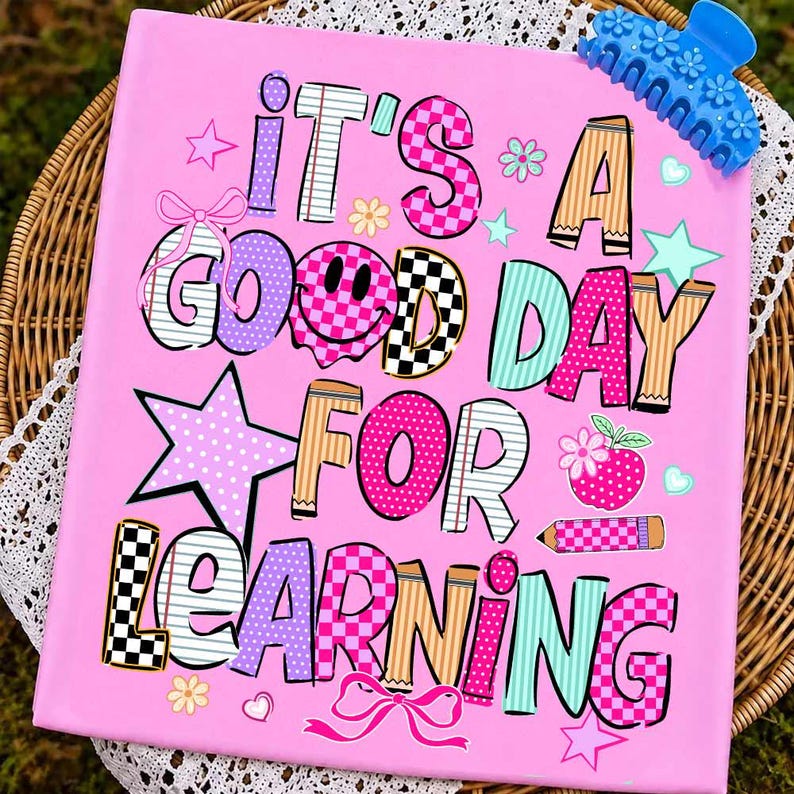 May include: A rectangular pink sign featuring the text IT'S A GOOD DAY FOR LEARNING in multi-colored, patterned letters. The design includes a smiley face, stars, bows, a pencil, and an apple icon. A blue floral hair claw clip rests on the top right.