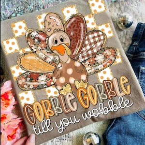 Puede incluir: Camiseta gris con un diseño de pavo de dibujos animados. El pavo tiene plumas estampadas y el texto "GOBBLE GOBBLE till you wobble" en naranja y marrón. El diseño incluye detalles de purpurina dorada y lunares.