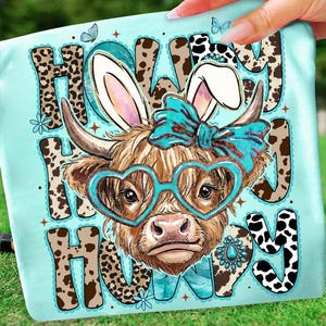 May include: A light blue graphic tee featuring a Highland cow with bunny ears, heart-shaped glasses, and a turquoise bow. The words "Howdy Hoppy" are printed in a mix of leopard print and cow spots.