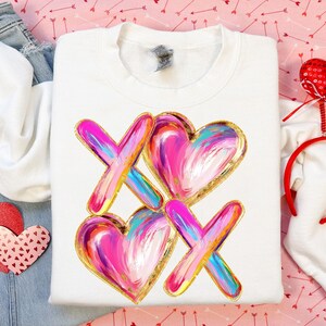 Puede incluir: Sudadera blanca con un dise&ntilde;o gr&aacute;fico de corazones y X rosas y dorados. Los corazones y las X est&aacute;n pintados con un efecto de pinceladas.