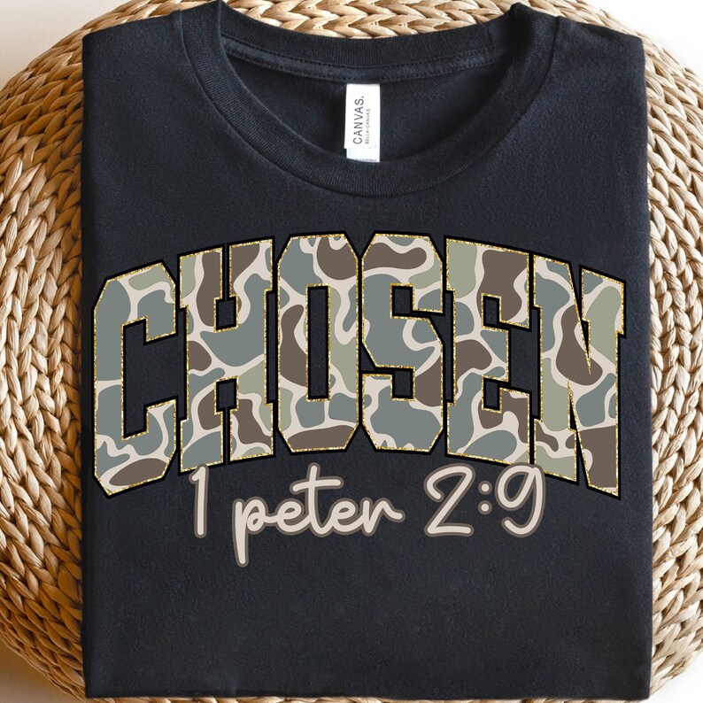 May include: Black t-shirt with the word "CHOSEN" in a camouflage pattern with gold trim. Below the word "CHOSEN" is the text "1 peter 2:9" in a light brown color. The t-shirt is displayed on a woven surface.