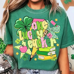 Può includere: T-shirt verde con la scritta "One Lucky Teacher" in lettere colorate e decorate. Il design include un quadrifoglio, una mela, una matita, libri e stelle, per la festa di San Patrizio e gli insegnanti.