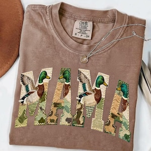 Peut inclure: Un t-shirt marron Comfort Colors avec le mot "MAMA" appliqué sur le devant. Chaque lettre présente un motif camouflage différent et une illustration de canard. Un collier argenté avec un pendentif est également visible.