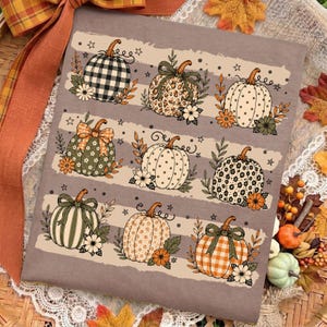 May include: A taupe t-shirt featuring a repeating pattern of nine pumpkins in three horizontal rows. The pumpkins display various prints including buffalo check, leopard spots, polka dots, and floral designs, accented with small stars and flowers.