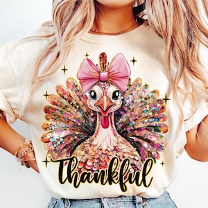 May include: A white t-shirt with a colorful glittery turkey wearing a pink bow and the word "thankful" in gold lettering.