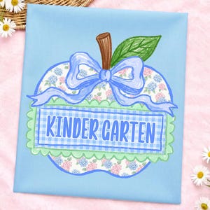 Op de afbeelding: Een lichtblauw stoffen vierkant met een centrale appelvormige afbeelding. De appel heeft een bloemenpatroon, een bruine steel, een groen blad en een lichtblauwe strik. Een rechthoekige blauwe gingham banner toont de tekst KINDER GARTEN.