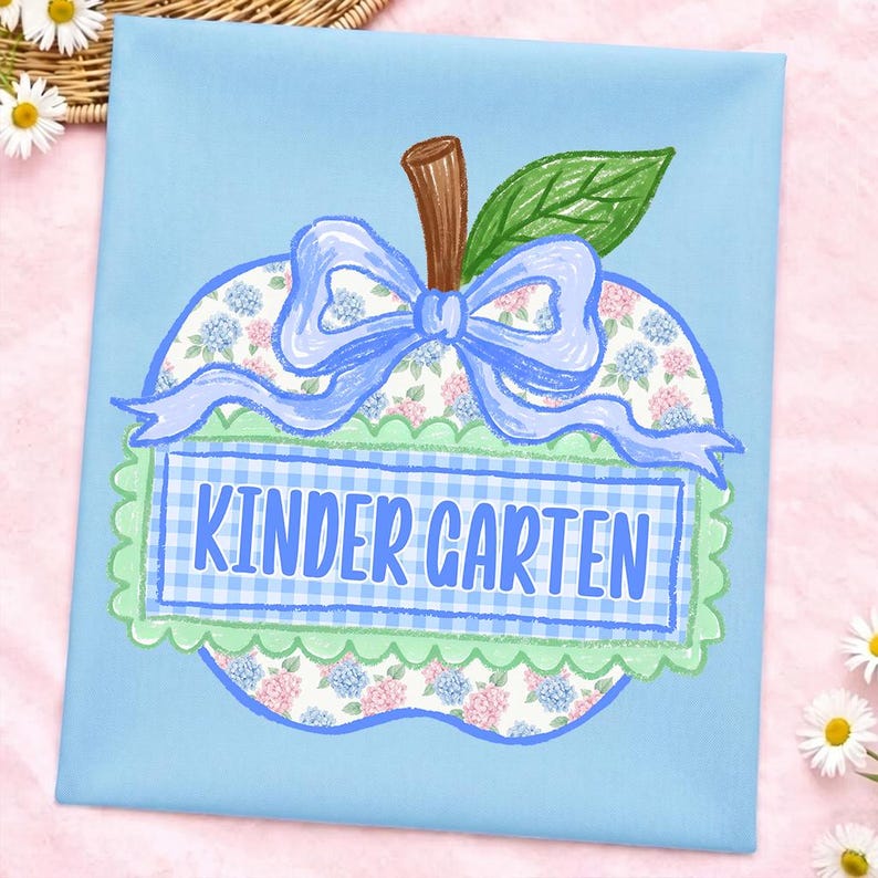 Op de afbeelding: Een lichtblauw stoffen vierkant met een centrale appelvormige afbeelding. De appel heeft een bloemenpatroon, een bruine steel, een groen blad en een lichtblauwe strik. Een rechthoekige blauwe gingham banner toont de tekst KINDER GARTEN.