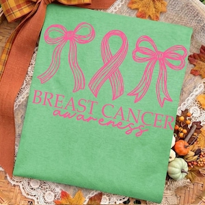 May include: A light green t-shirt featuring a graphic of two pink bows flanking a pink awareness ribbon. Below the graphics, the text "BREAST CANCER awareness" is printed in pink. The design is centered on the front of the fabric.