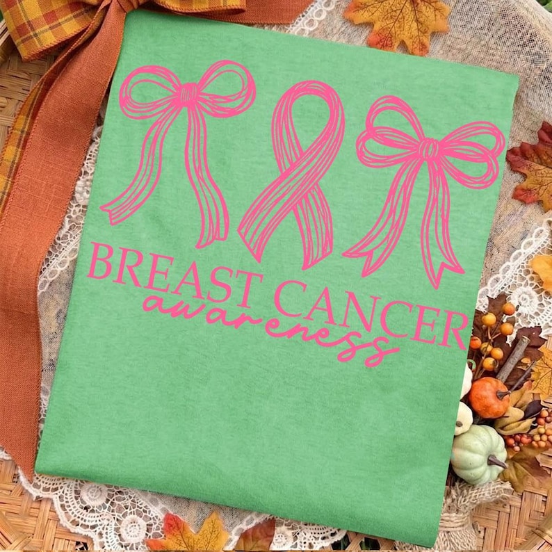 May include: A light green t-shirt featuring a graphic of two pink bows flanking a pink awareness ribbon. Below the graphics, the text "BREAST CANCER awareness" is printed in pink. The design is centered on the front of the fabric.