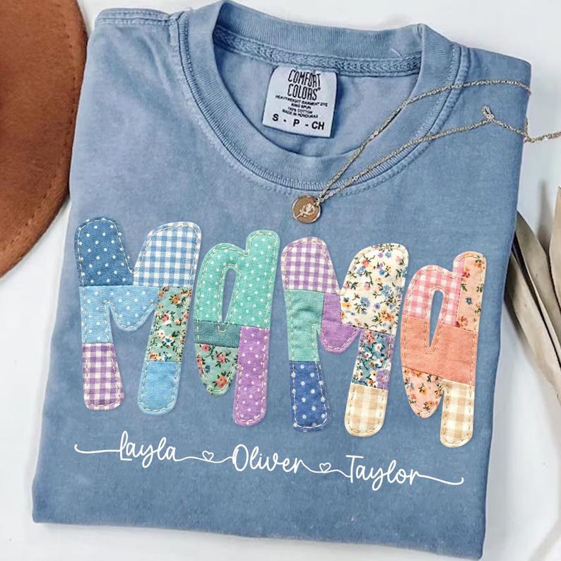 May include: Blue Comfort Colors t-shirt with the word "Mama" in patchwork letters. The letters are various colors and patterns. Below the word "Mama" are the names "Layla", "Oliver", and "Taylor". A gold necklace rests on the shirt.