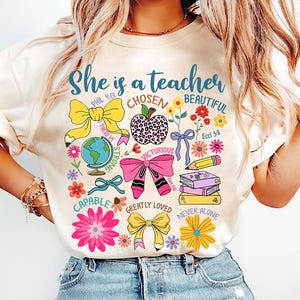 以下が含まれることがあります： 「She is a teacher」というテキストと、「Chosen」、「Beautiful」、「Strong」、「Victorious」、「Capable」、「Greatly Loved」、「Never Alone」などのさまざまなインスピレーションを与える言葉やフレーズが描かれたカラフルなデザインの白いTシャツ。
