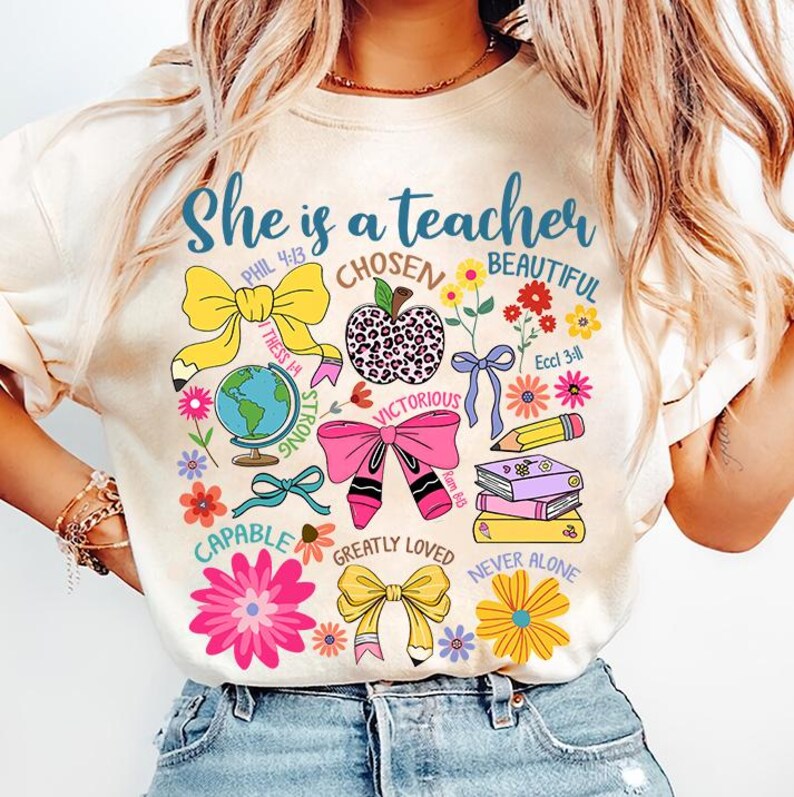 以下が含まれることがあります： 「She is a teacher」というテキストと、「Chosen」、「Beautiful」、「Strong」、「Victorious」、「Capable」、「Greatly Loved」、「Never Alone」などのさまざまなインスピレーションを与える言葉やフレーズが描かれたカラフルなデザインの白いTシャツ。