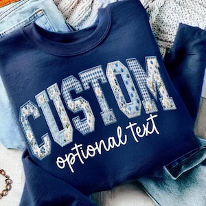 Może przedstawiać: Granatowa bluza z napisem "CUSTOM" w patchworkowym wzorze z kwiatowymi, kratkowanymi i groszkowymi wzorami. Słowa "optional text" są napisane poniżej białym skryptem. Bluza jest złożona na niebieskich dżinsach.