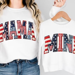May include: White sweatshirts with patriotic "MAMA" and "MINI" text. The letters are designed with red and white stripes, blue stars, and a red and white checkered pattern, perfect for matching outfits.
