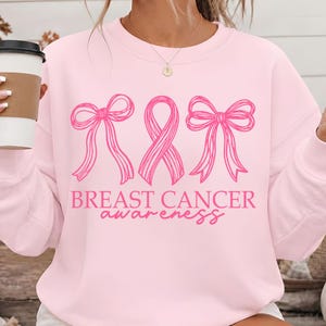 May include: A light pink crewneck sweatshirt featuring a dark pink graphic print. The design includes two bows flanking a central awareness ribbon, with the text "BREAST CANCER awareness" printed in a combination of block and script fonts.