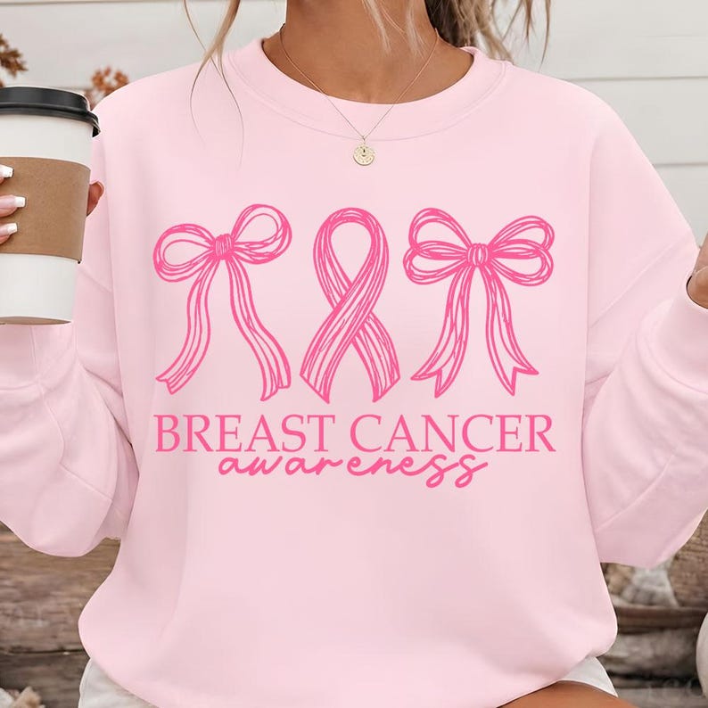 May include: A light pink crewneck sweatshirt featuring a dark pink graphic print. The design includes two bows flanking a central awareness ribbon, with the text "BREAST CANCER awareness" printed in a combination of block and script fonts.