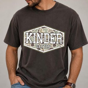 May include: Dark grey t-shirt featuring a camouflage design within a white-bordered hexagon. The text "KINDER GARTEN" is displayed in a distressed, white font. The shirt is a casual, short-sleeved style.