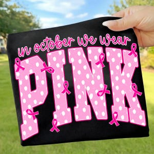 Puede incluir: Una camiseta negra con el texto "in october we wear" en letra rosa sobre la palabra "PINK" en letras grandes, rosas y con lunares blancos. Varias cintas rosas peque&ntilde;as de concienciaci&oacute;n sobre el c&aacute;ncer de mama est&aacute;n sobre las letras.