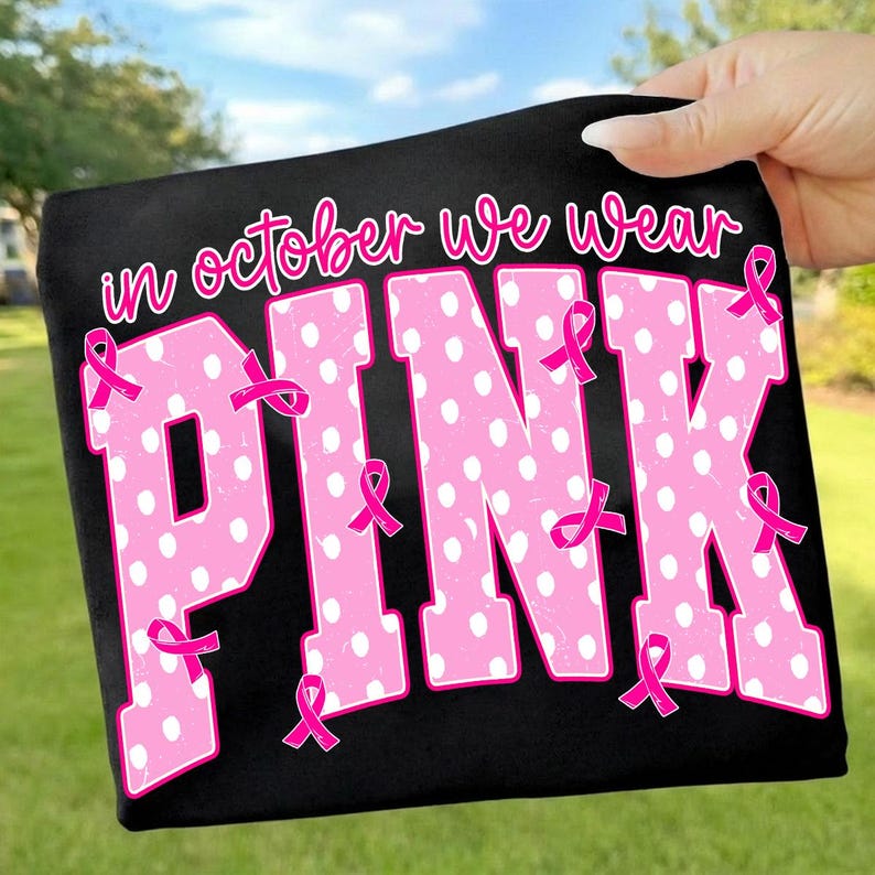Puede incluir: Una camiseta negra con el texto "in october we wear" en letra rosa sobre la palabra "PINK" en letras grandes, rosas y con lunares blancos. Varias cintas rosas peque&ntilde;as de concienciaci&oacute;n sobre el c&aacute;ncer de mama est&aacute;n sobre las letras.