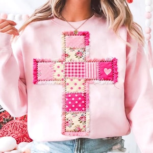 May include: A light pink sweatshirt featuring a patchwork cross design. The cross is made of various pink fabrics, including floral, polka dot, and striped patterns, with a heart detail. The edges of the cross have a frayed trim.