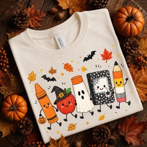 May include: A cream-colored t-shirt featuring a graphic print of five anthropomorphic school supplies and an apple dressed for Halloween. The characters include an orange crayon, a red apple with a witch hat, a white ghost, a black notebook, and a pencil.