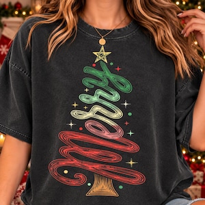 May include: A charcoal grey short-sleeve t-shirt featuring a graphic of a Christmas tree formed by the word Merry in cursive script. The tree is coloured in green, yellow, and red, topped with a yellow star, and accented with small star and dot icons.