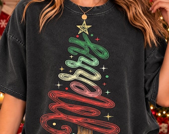May include: A charcoal gray short-sleeve t-shirt featuring a graphic of a Christmas tree formed by the word Merry in cursive script. The tree is colored in green, yellow, and red, topped with a yellow star, and accented with small star and dot icons.