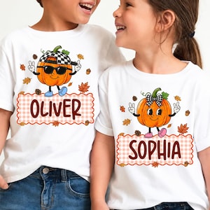 May include: Two white t-shirts featuring a cartoon pumpkin graphic. The left shirt displays a pumpkin wearing a checkered cap and sunglasses with the name OLIVER. The right shirt shows a pumpkin with a checkered bow and the name SOPHIA.