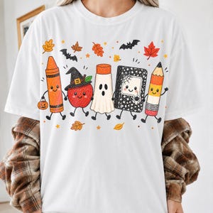 May include: A white t-shirt featuring a graphic print of five anthropomorphic school supplies including an orange crayon, a red apple wearing a witch hat, a white ghost, a black composition notebook, and a yellow pencil, surrounded by autumn leaves.