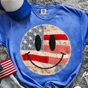 May include: A blue t-shirt featuring a smiley face design with an American flag motif. The smiley face is made of denim, red and white patterned fabric, and black oval eyes. A blue and white trucker hat is visible in the background.