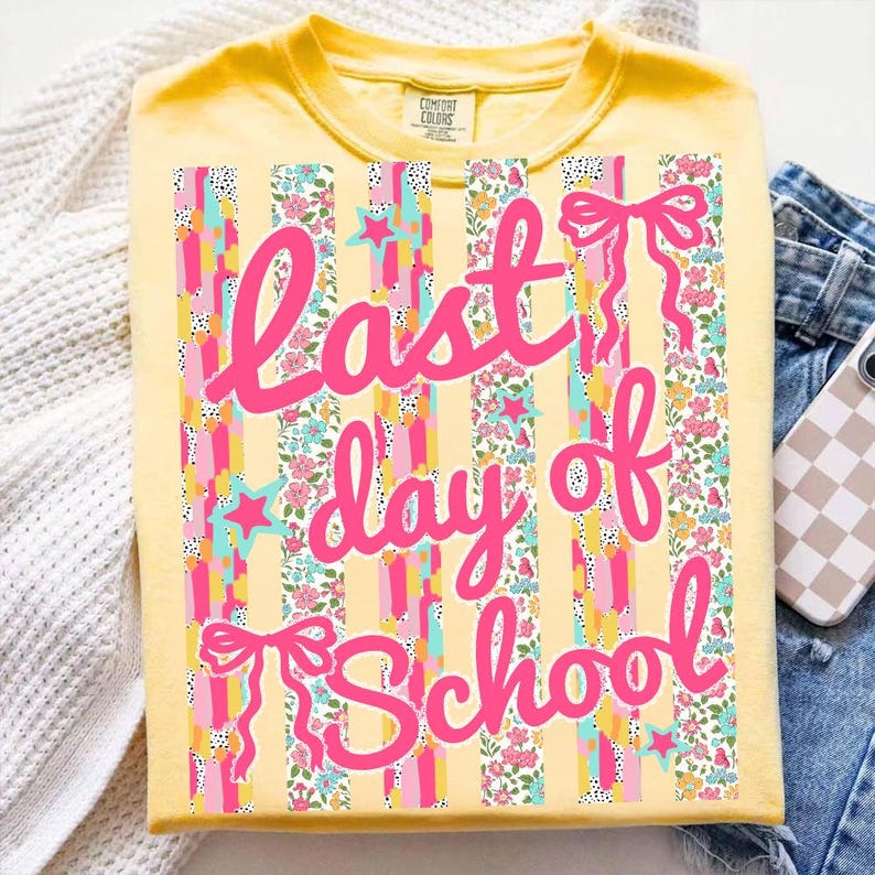 Puede incluir: Camiseta amarilla con la frase "Last day of School" en rosa. El dise&ntilde;o incluye rayas verticales, estampados florales, estrellas y lazos. La camiseta est&aacute; doblada, con vaqueros y una funda de tel&eacute;fono en el fondo.