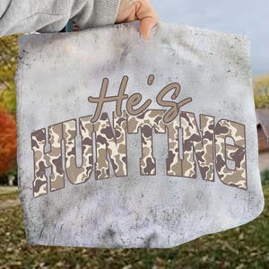 May include: A gray tote bag with the text "He's HUNTING". The word "HUNTING" is in a camouflage pattern. The bag is held by a person's hand.