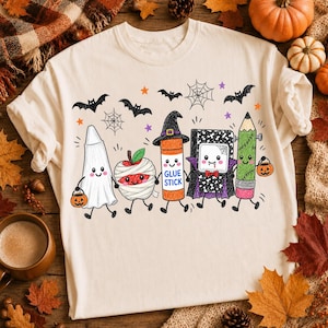 Pode incluir: Uma t-shirt cor de creme com um gr&aacute;fico colorido de cinco materiais escolares antropom&oacute;rficos vestidos para o Halloween: um fantasma, uma ma&ccedil;&atilde; m&uacute;mia, uma cola em bast&atilde;o de bruxa, um caderno vampiro e um l&aacute;pis Frankenstein, com morcegos.