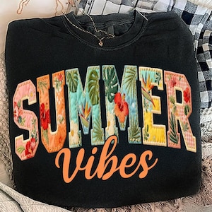May include: Black sweatshirt with the words "SUMMER vibes" in a colorful, floral design. The letters are filled with various tropical flowers and leaves in shades of pink, orange, green, and yellow. A gold necklace is visible.