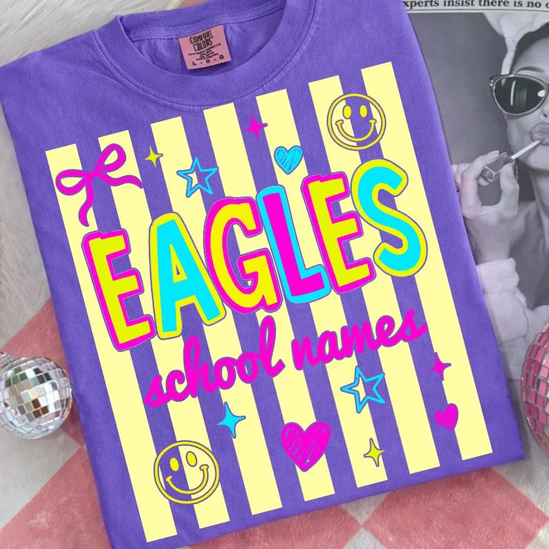 Puede incluir: Camiseta morada con un dise&ntilde;o de rayas amarillas y blancas. La palabra "EAGLES" est&aacute; en letras grandes y coloridas, con "school names" debajo. La camiseta tiene caras sonrientes, estrellas, corazones y un lazo.