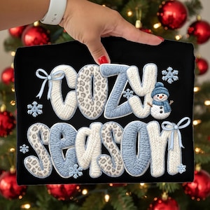 Pu&ograve; includere: T-shirt nero con la scritta "COZY SEASON" in stile maglia. Le lettere sono bianche, blu e con stampa leopardata. Include fiocchi di neve e un pupazzo di neve con cappello e sciarpa blu.