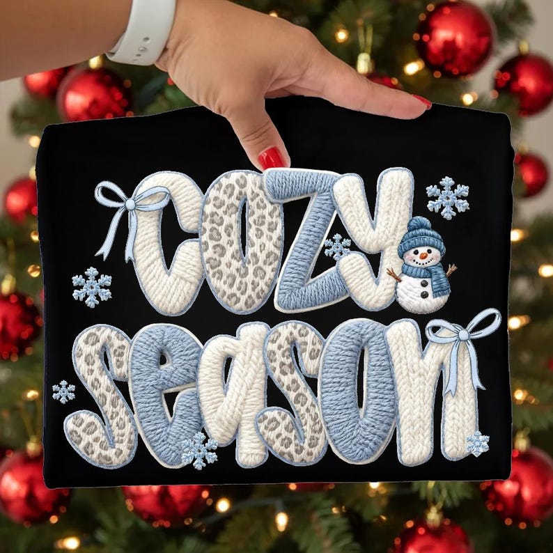 Pu&ograve; includere: T-shirt nero con la scritta "COZY SEASON" in stile maglia. Le lettere sono bianche, blu e con stampa leopardata. Include fiocchi di neve e un pupazzo di neve con cappello e sciarpa blu.