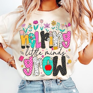 May include: A white t-shirt with a colorful graphic that says "helping little minds grow". The graphic features flowers, pencils, a lightbulb, and a red apple.