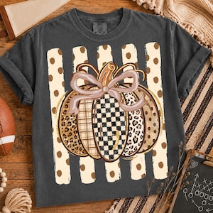 K&ouml;nnte beinhalten: Ein anthrazitfarbenes Kurzarm-T-Shirt mit einer zentralen Grafik eines K&uuml;rbisses mit einer hellrosa Schleife. Der K&uuml;rbis besteht aus Segmenten mit Leopardenmuster, Karomuster und Schachbrettmuster vor vertikalen cremefarbenen Streifen.