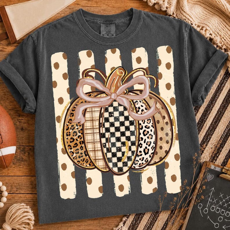 K&ouml;nnte beinhalten: Ein anthrazitfarbenes Kurzarm-T-Shirt mit einer zentralen Grafik eines K&uuml;rbisses mit einer hellrosa Schleife. Der K&uuml;rbis besteht aus Segmenten mit Leopardenmuster, Karomuster und Schachbrettmuster vor vertikalen cremefarbenen Streifen.