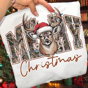 May include: White t-shirt featuring a deer wearing a Santa hat and Christmas lights. The word "MERRY" is printed in a camouflage pattern, and "Christmas" is written in a brown script font. A festive holiday design.