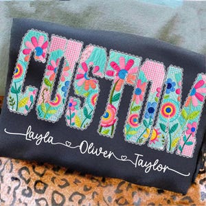 May include: Dark gray sweatshirt with the word "CUSTOM" in colorful floral patchwork letters. Below the word are the names "Layla", "Oliver", and "Taylor" in white script.