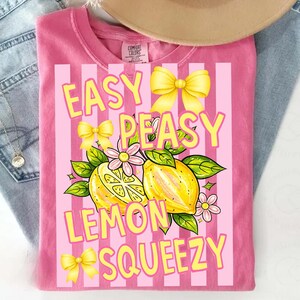May include: Pink t-shirt with a graphic design featuring the text "EASY PEASY LEMON SQUEEZY" in yellow. The design includes yellow bows, lemons, and flowers. The shirt is folded on a light blue denim surface.