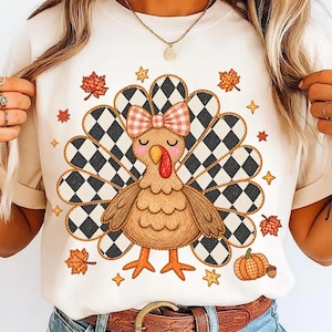 May include: A cream-colored t-shirt featuring a central graphic of a turkey with a black and white checkered tail, a pink and white gingham bow on its head, and a red wattle. The design is surrounded by small orange autumn leaves and stars.