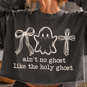May include: A charcoal grey long-sleeve sweatshirt featuring a cream-coloured graphic print of a bow, a smiling ghost, and a cross. Below the graphics, the text reads: ain't no ghost like the holy ghost. The garment has a raw, unfinished hemline.
